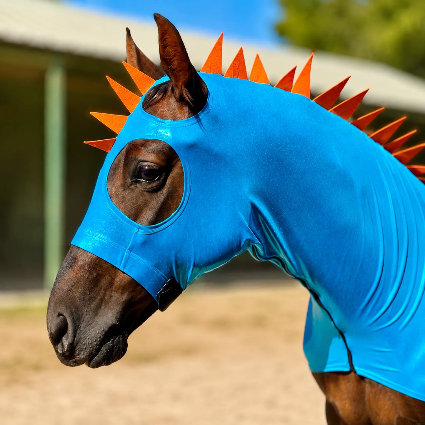 Horse wearing a metallic blue dragon horse costume with orange spikes and a matching tailbag at an outdoor setting.