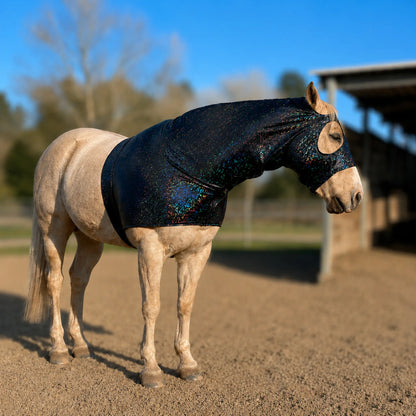 A light-colored horse standing in an outdoor stable arena wearing a dark iridescent hologram black sleazy hood showing the right side of the horse.