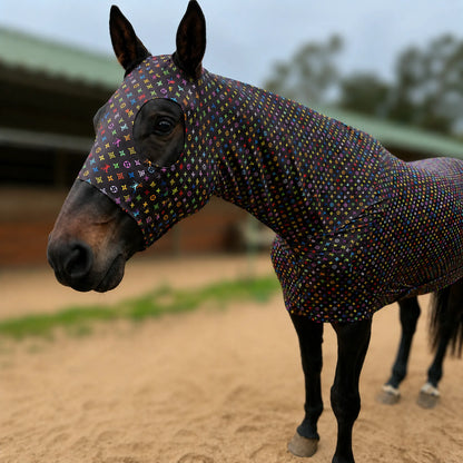A horse wearing a fitted LV color full body sleazy, photographed outdoors in a sandy paddock with a stable in the background.