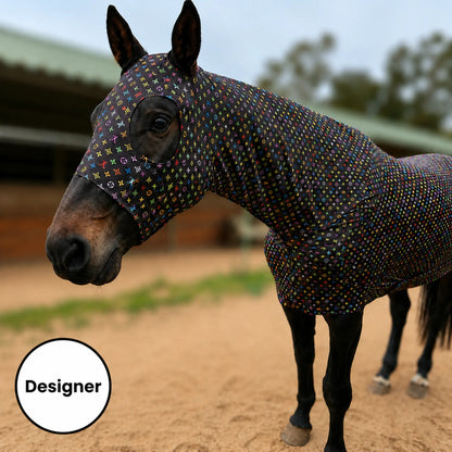 A horse wearing a fitted LV color full body, photographed outdoors in a sandy arena with a stable in the background.