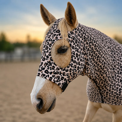 A horse wearing a leopard-print horse hood, standing in a sandy outdoor arena at sunset.