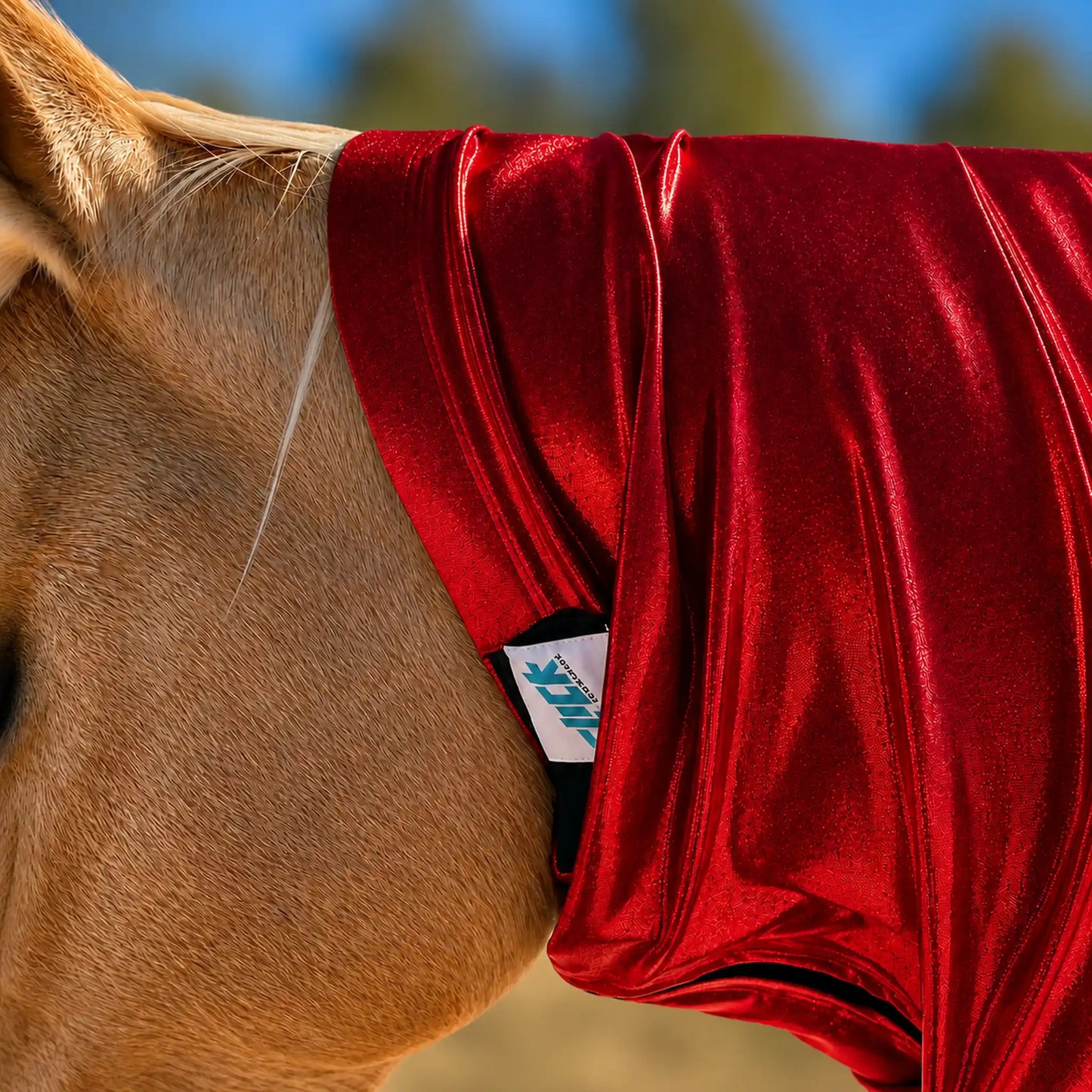 A close-up of a horse wearing a shiny metallic red faceless sleazy hood, with a small visible Slick Horseware brand tag near the neck area. The horse's tan coat and part of its ear are visible against a softly blurred outdoor background.
