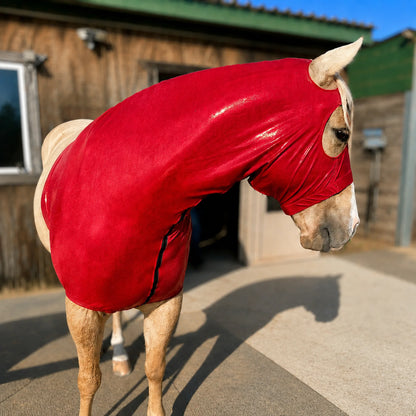 A light-colored horse standing outdoors in front of a wooden stable, wearing a bright metallic red sleazy hood with eye and nose openings.
