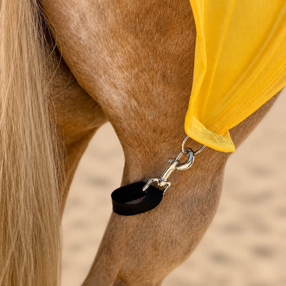 Close-up of a horse's leg strap with metal hardware with a bright yellow full body sleazy cover, set against a blurred sandy background.