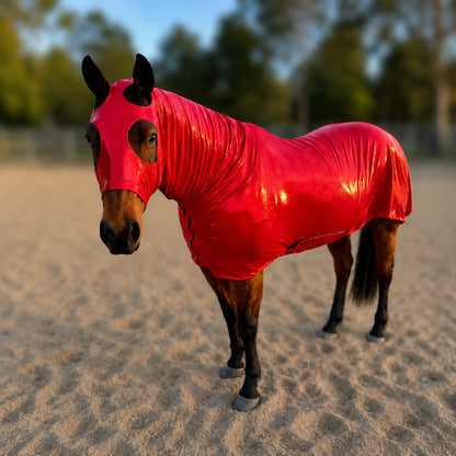 A glossy metallic red full body horse sleazy worn by a brown horse standing in a sandy outdoor arena with blurred trees in the background.