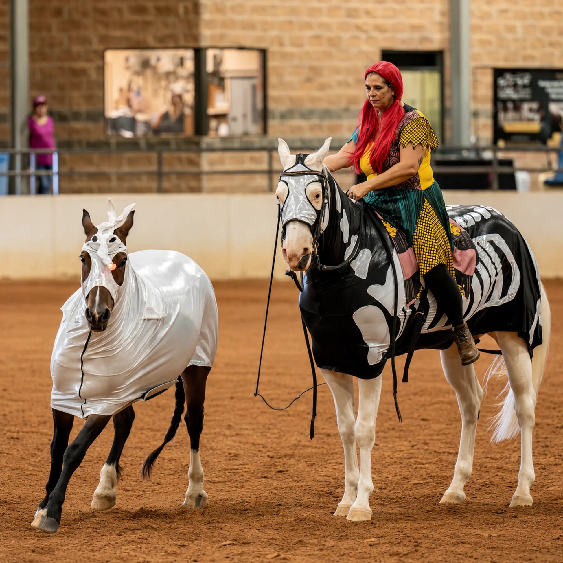 A woman in colorful clothing rides a white horse wearing a skeleton patterned sleazy inside an indoor arena, while a second white horse beside it wears a metallic silver full body sleazy costume.