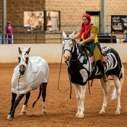 A woman in colorful clothing rides a white horse wearing a skeleton patterned sleazy inside an indoor arena, while a second white horse beside it wears a metallic silver full body sleazy costume.