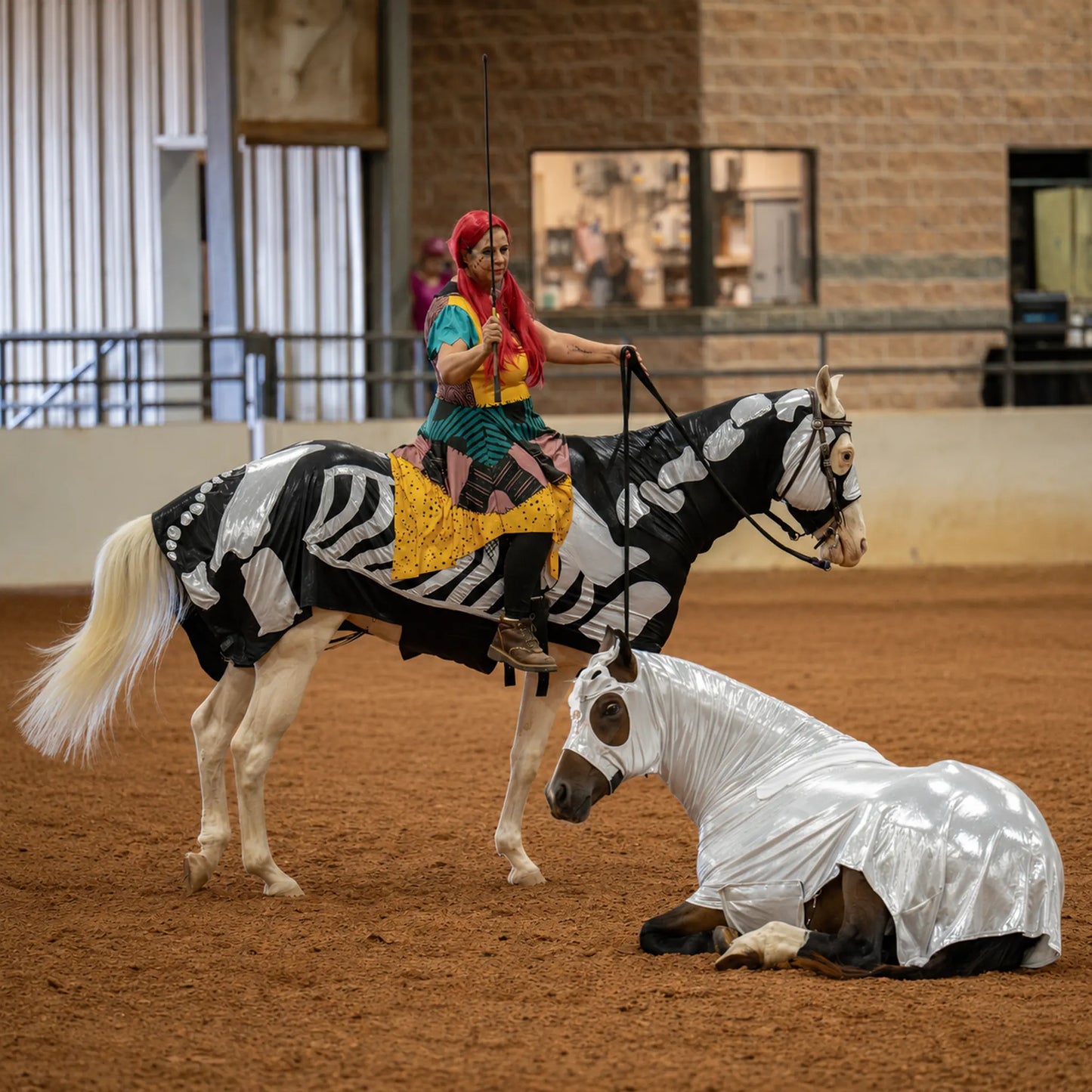 A person in colorful costume rides a white horse with a black-and-white skeleton print while holding a sword, with another metallic silver full body sleazy costume on a horse lying on the reddish-brown dirt.