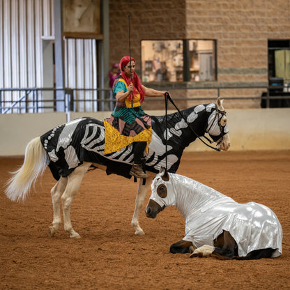 A person in colorful costume rides a white horse with a black-and-white skeleton print while holding a sword, with another metallic silver full body sleazy costume on a horse lying on the reddish-brown dirt.
